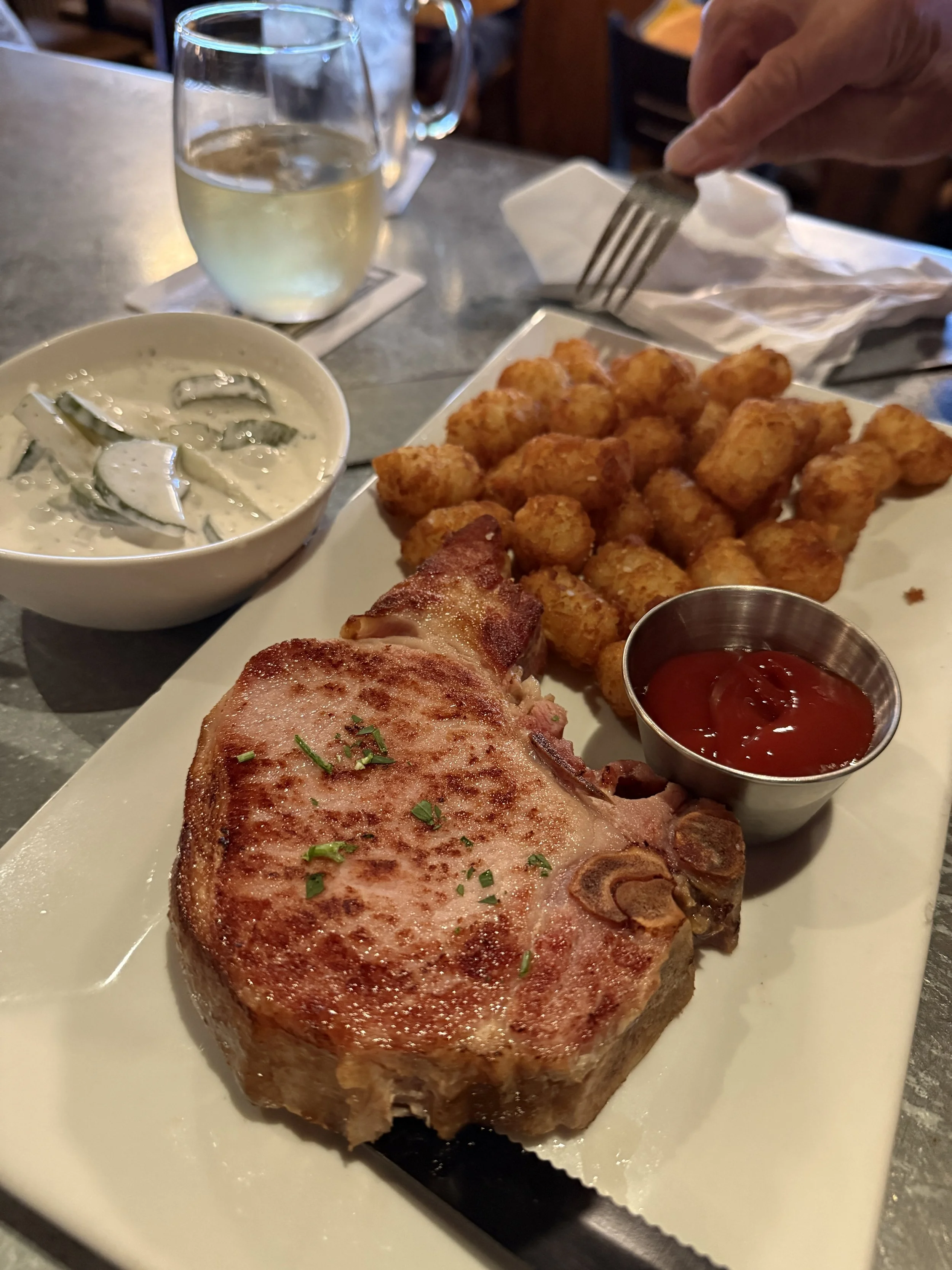 A plate with a large, cooked pork chop garnished with chopped herbs, alongside a side of fried cheese curds, a cup of ketchup, and a small bowl of creamy cucumber salad, with a glass of white wine in the background.