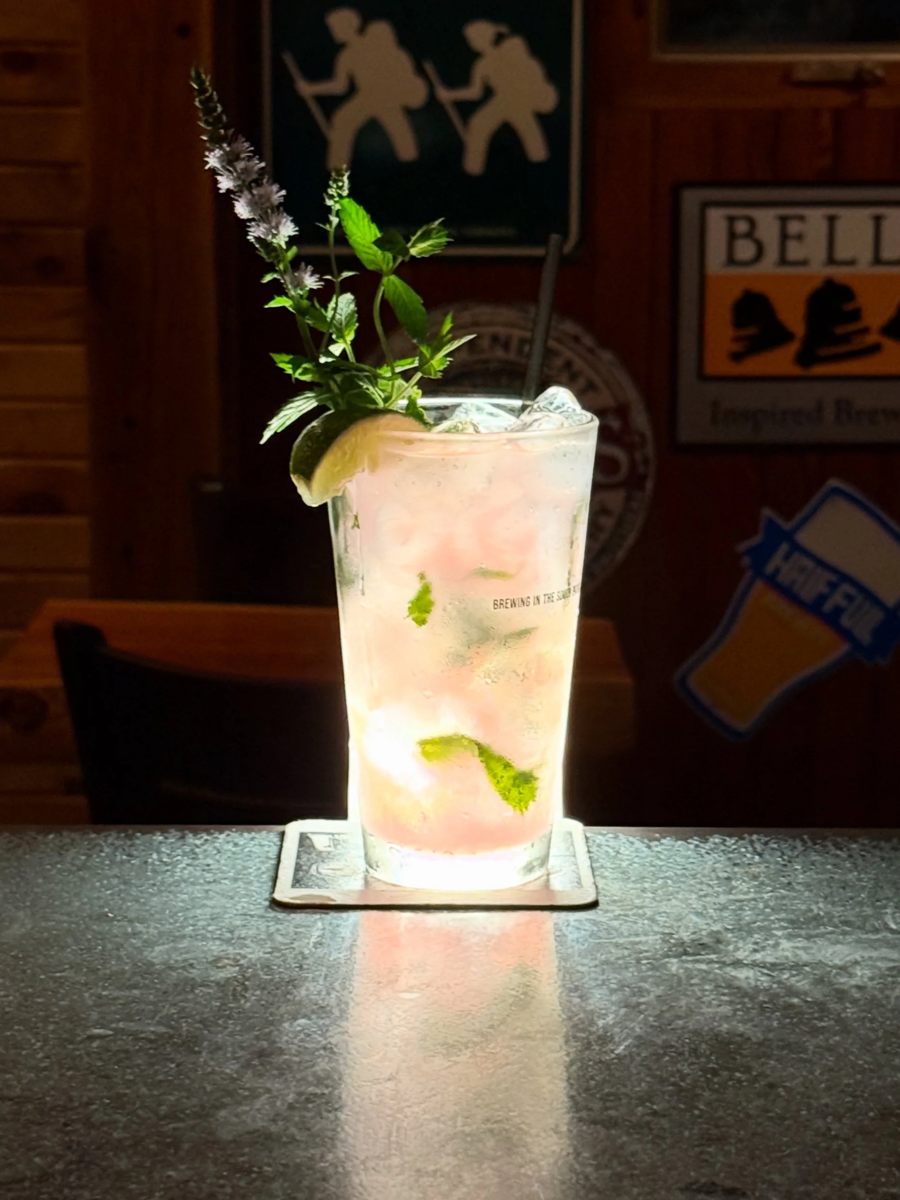 A cocktail glass with ice, mint, and a lemon wedge, illuminated from beneath, with a dark wooden wall and patterned decorations in the background.