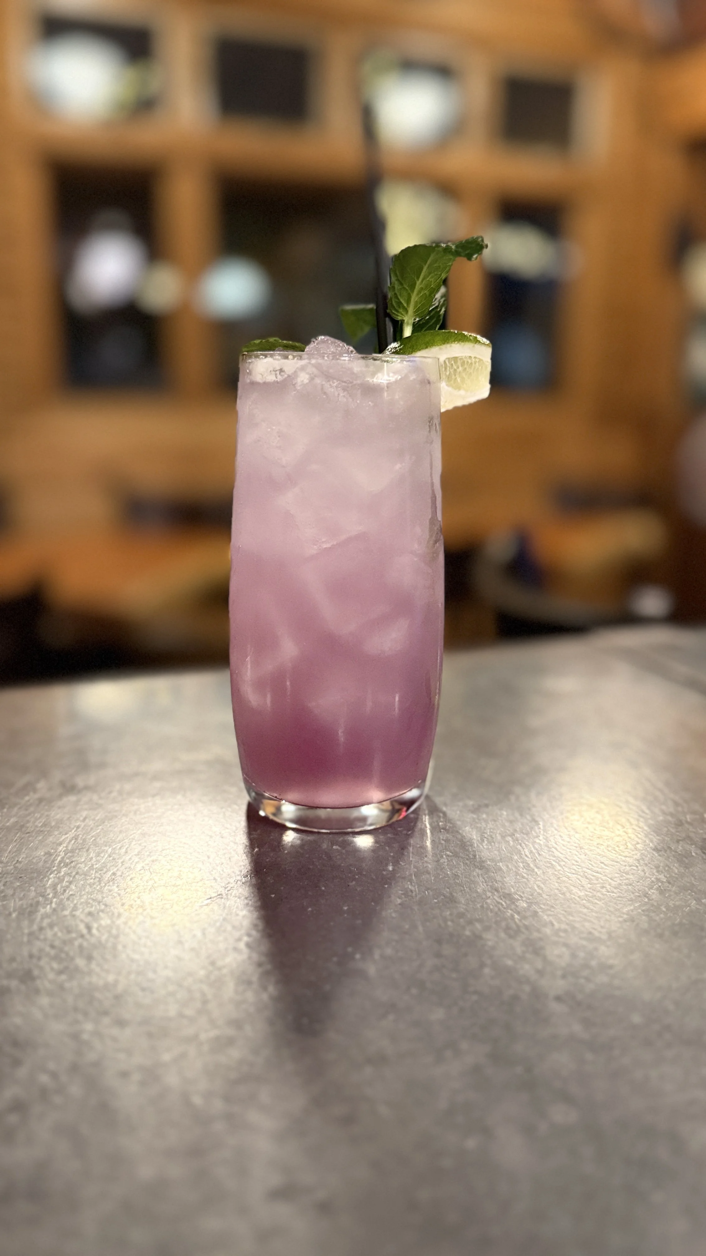 A tall glass of pink cocktail with ice, garnished with lime wedges and fresh mint, on a dark surface with a wooden cubical display in the background.