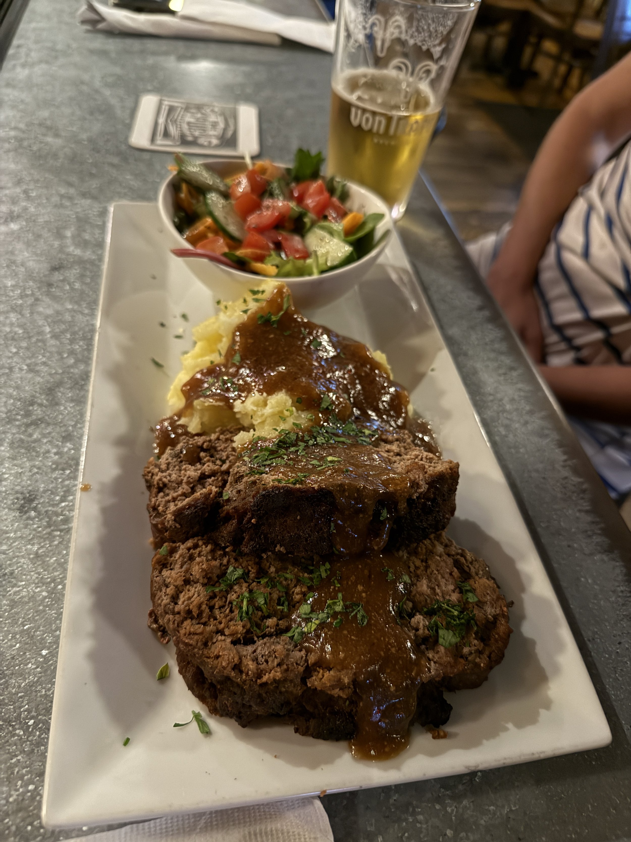 A white rectangular plate with two pieces of meatloaf topped with brown gravy, garnished with chopped herbs. Next to it is a bowl of mixed fresh salad including tomatoes, cucumbers, and greens. In the background, there is a tall glass of beer on a ta