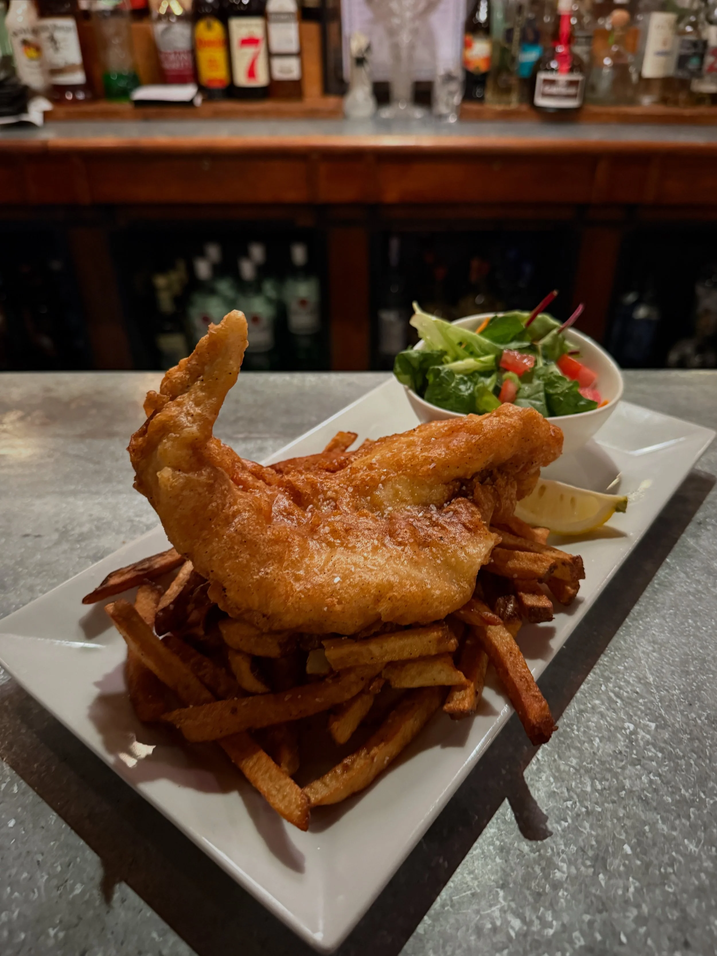 Fried fish served on a bed of French fries with a side salad of spinach, tomatoes, and a lemon wedge.