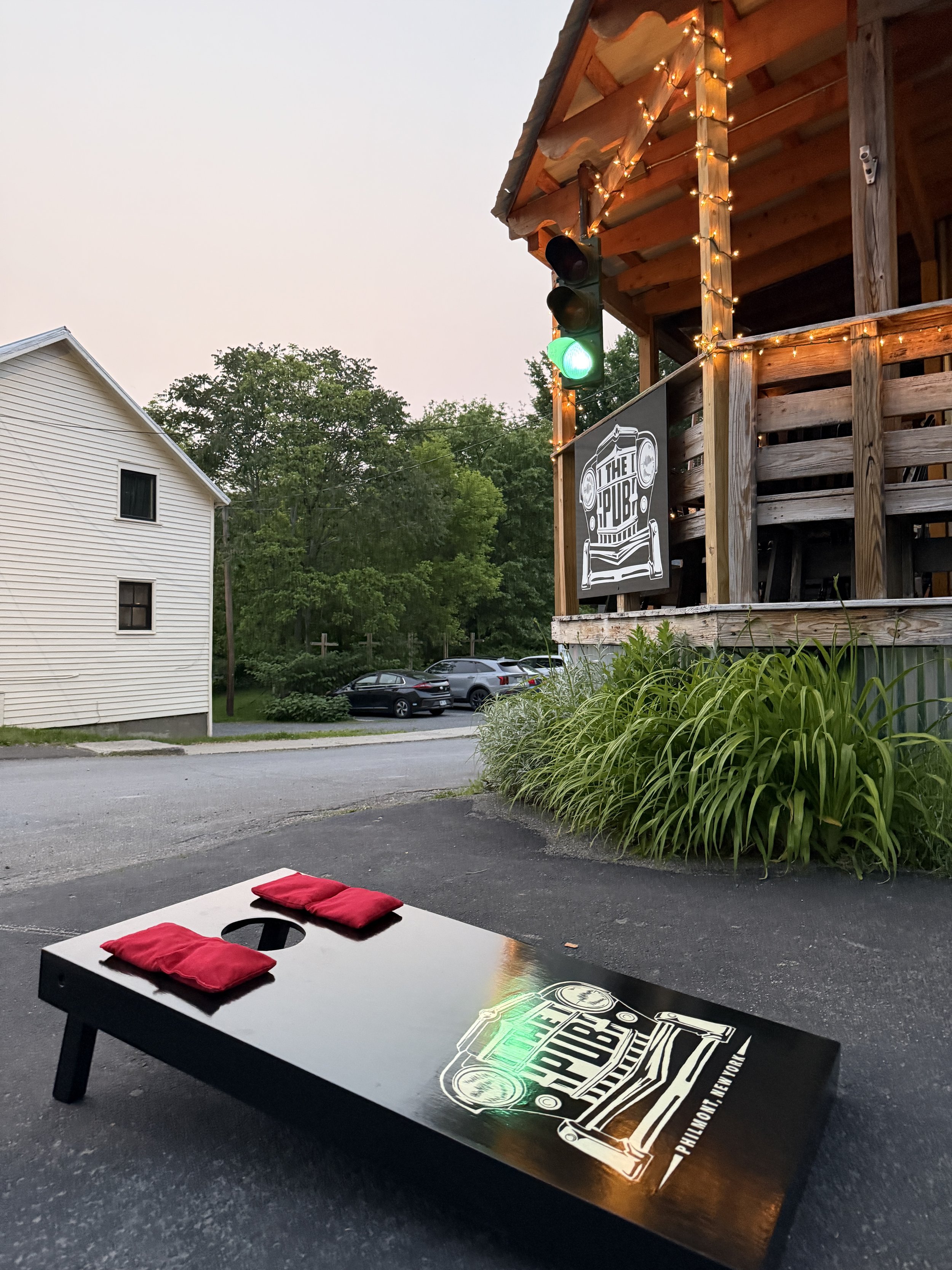 An outdoor cornhole game with a black board and red bags, set up on a paved area near a pub. In the background, a wooden building with string lights and a green traffic light are visible, along with trees and parked cars.