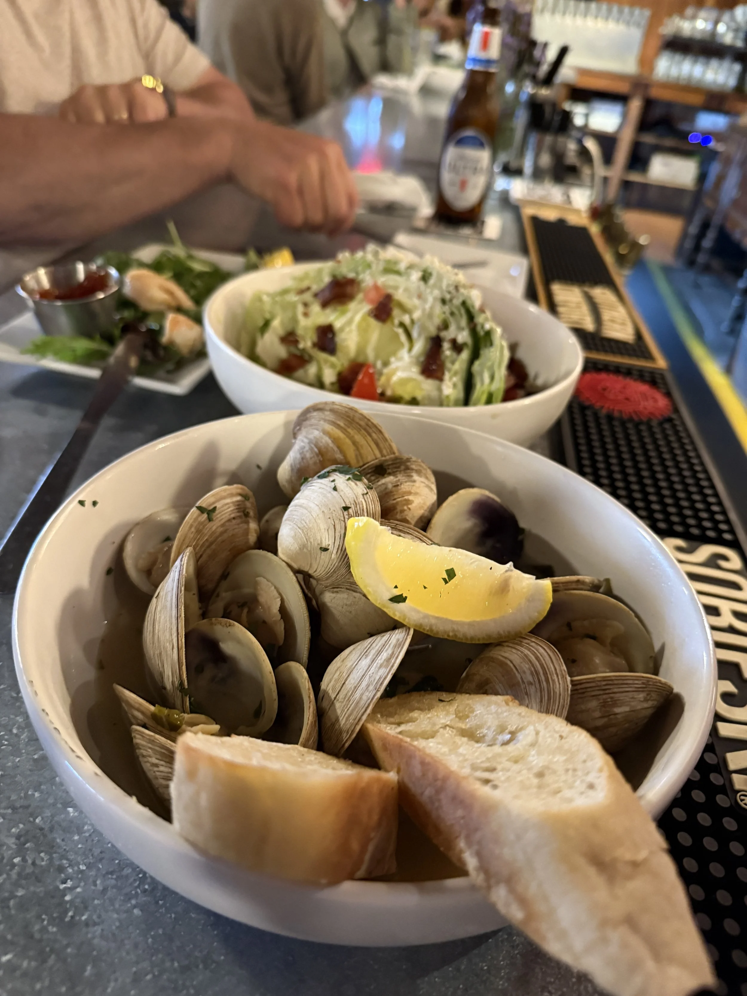 A bowl of cooked clams with lemon wedge and herbs, and slices of bread in the foreground. In the background, a salad, a bottle of beer, and a person with folded arms.