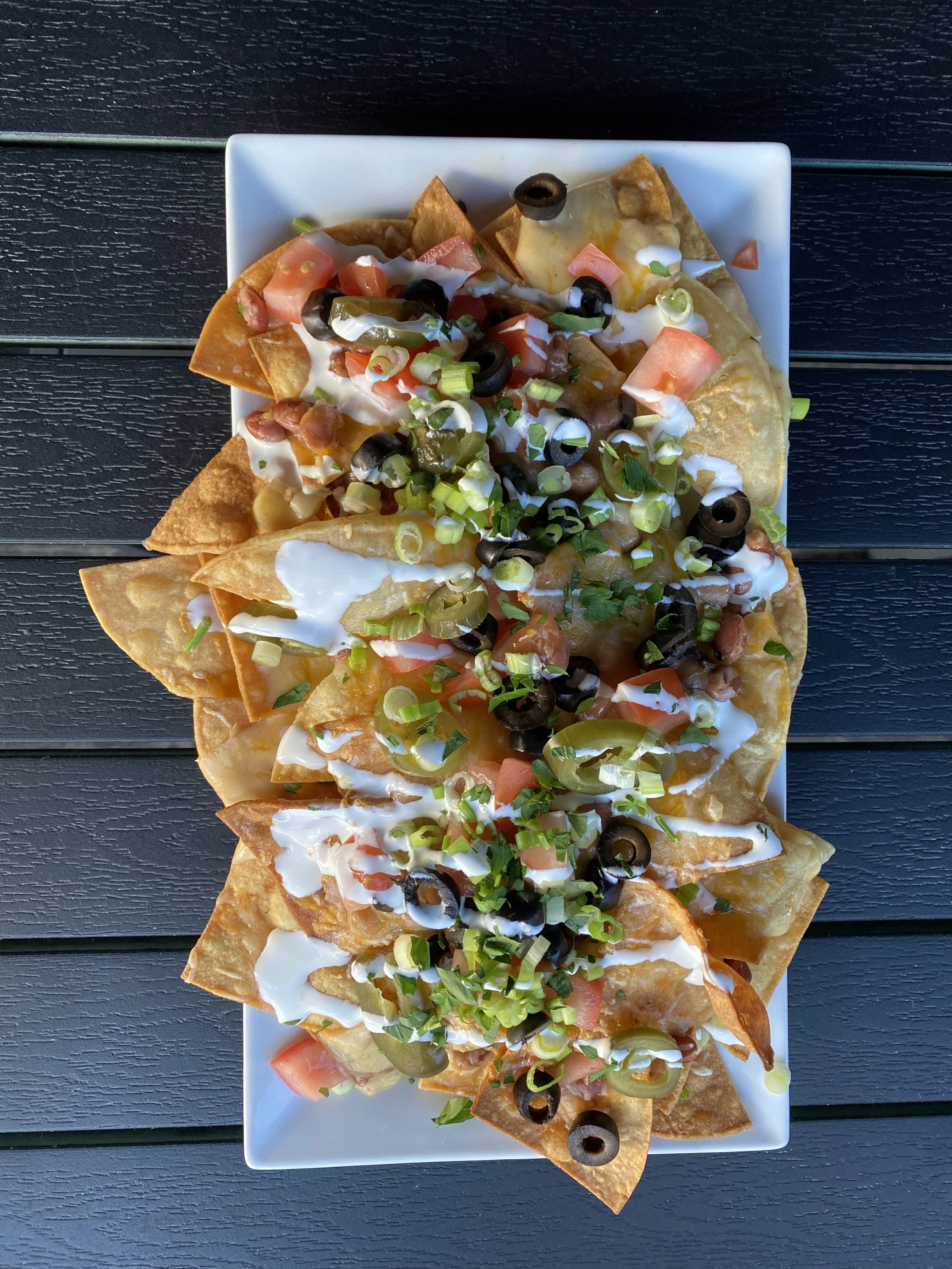 A plate of nachos topped with black olives, chopped green onions, diced tomatoes, sour cream, and melted cheese on a black wooden table.