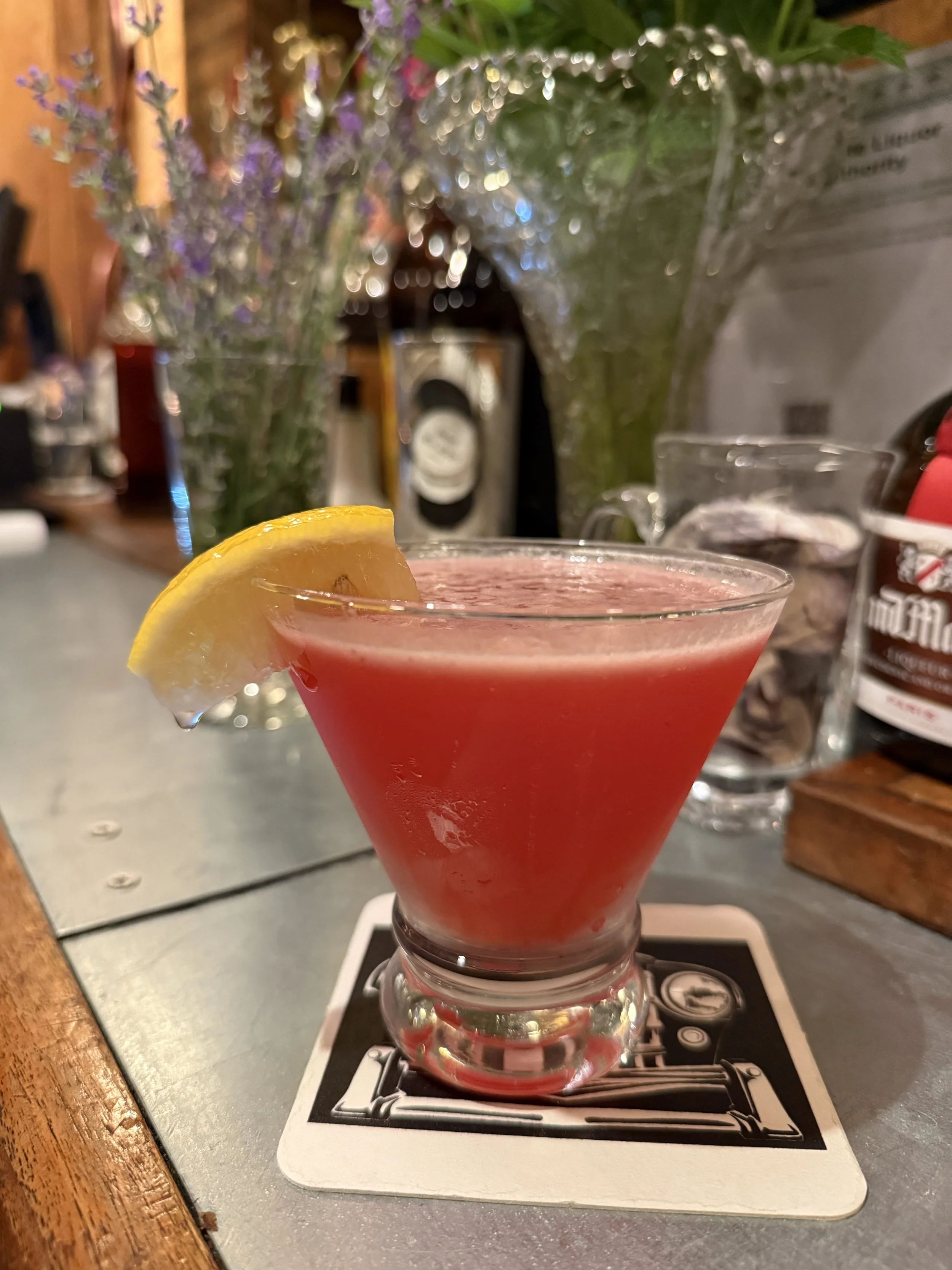 A red cocktail with a lemon wedge garnish on a bartender's coaster. Blurred background with flowers and bottles.