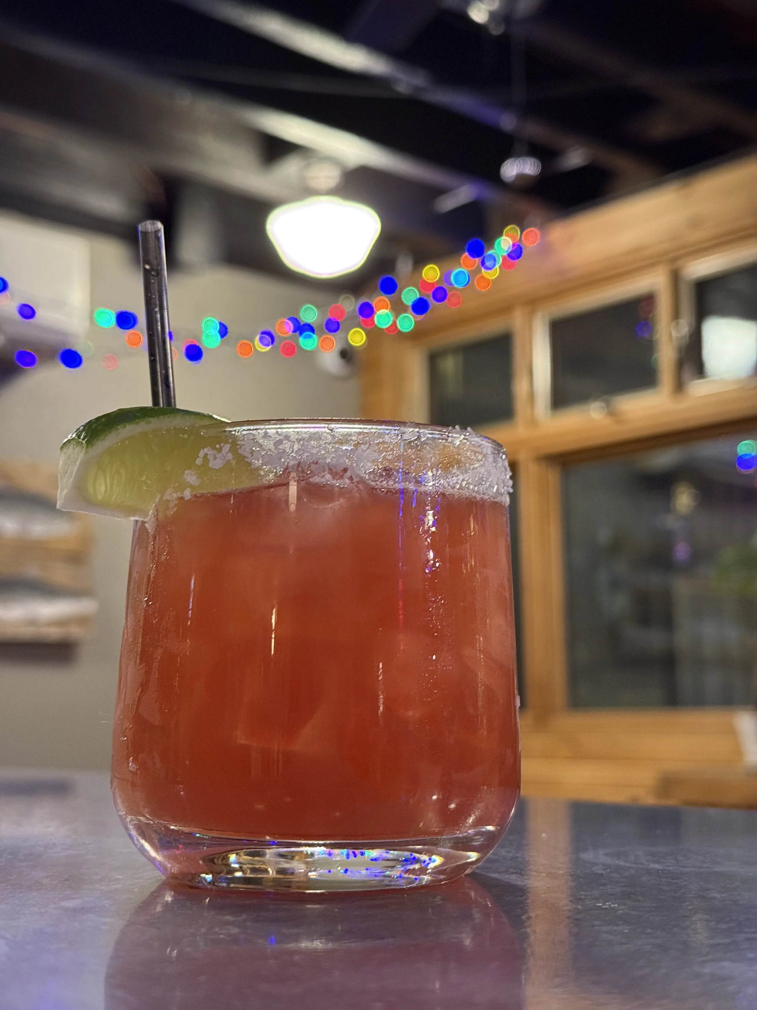 Close-up of a pink cocktail with a lime wedge garnish and a black straw in a clear glass, with colorful blurry Christmas lights hanging in the background.