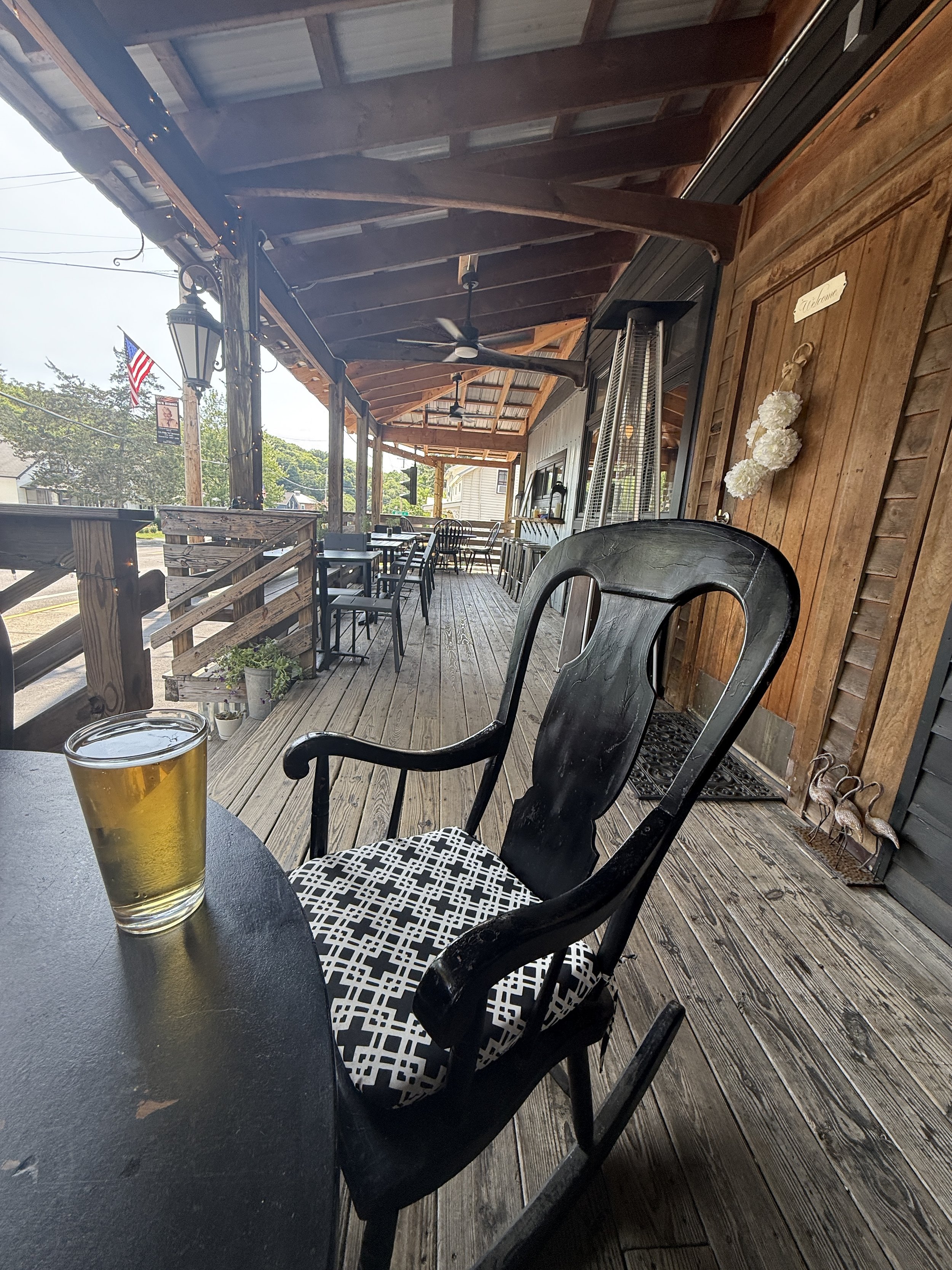 Outdoor restaurant patio with wooden flooring, black chairs, and a table with a pint of beer. There are potted plants, a wooden board with white flowers, a fan, and a heater. An American flag is visible in the background.