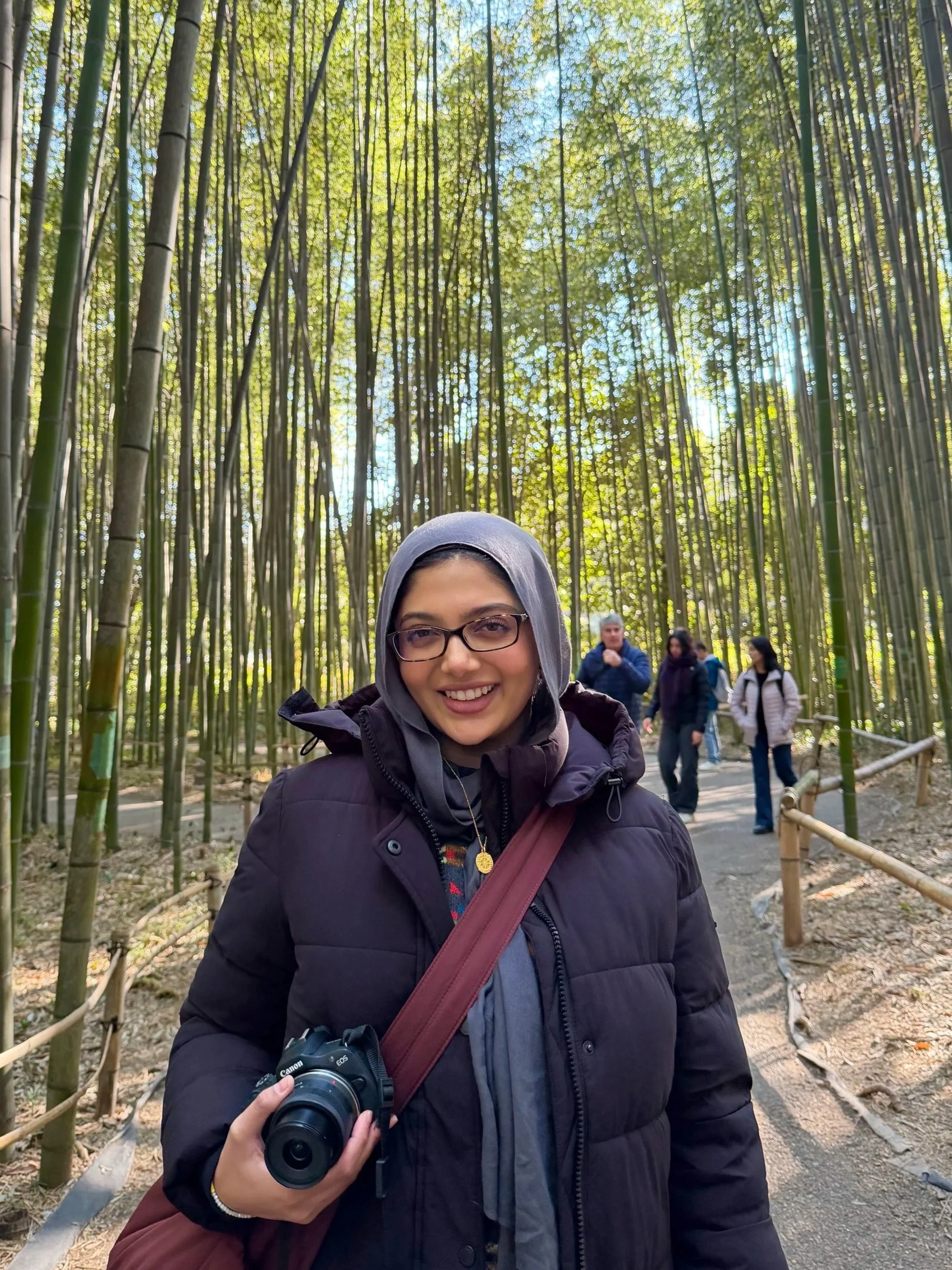 A woman wearing glasses and a hoodie smiling at the camera, holding a camera in her right hand, standing in a bamboo forest with a group of people in the background.