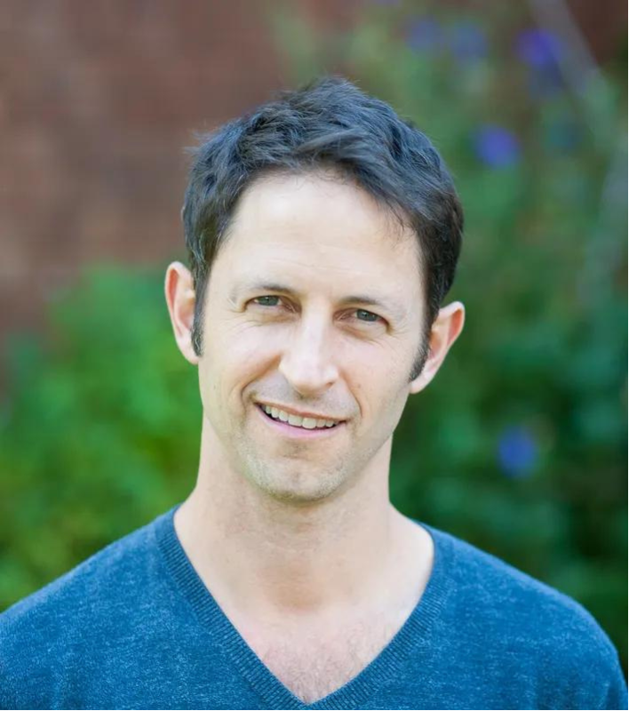 A man with short dark hair, light skin, and blue eyes, smiling outdoors with a blurred green background.