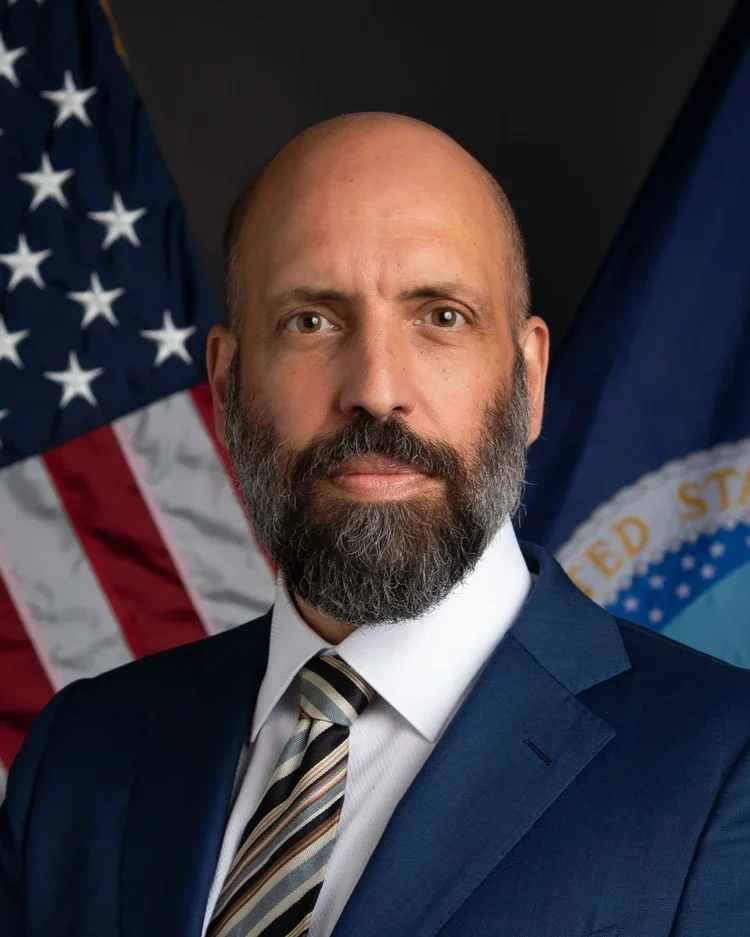 A man with a beard in a suit and tie standing in front of American flags.