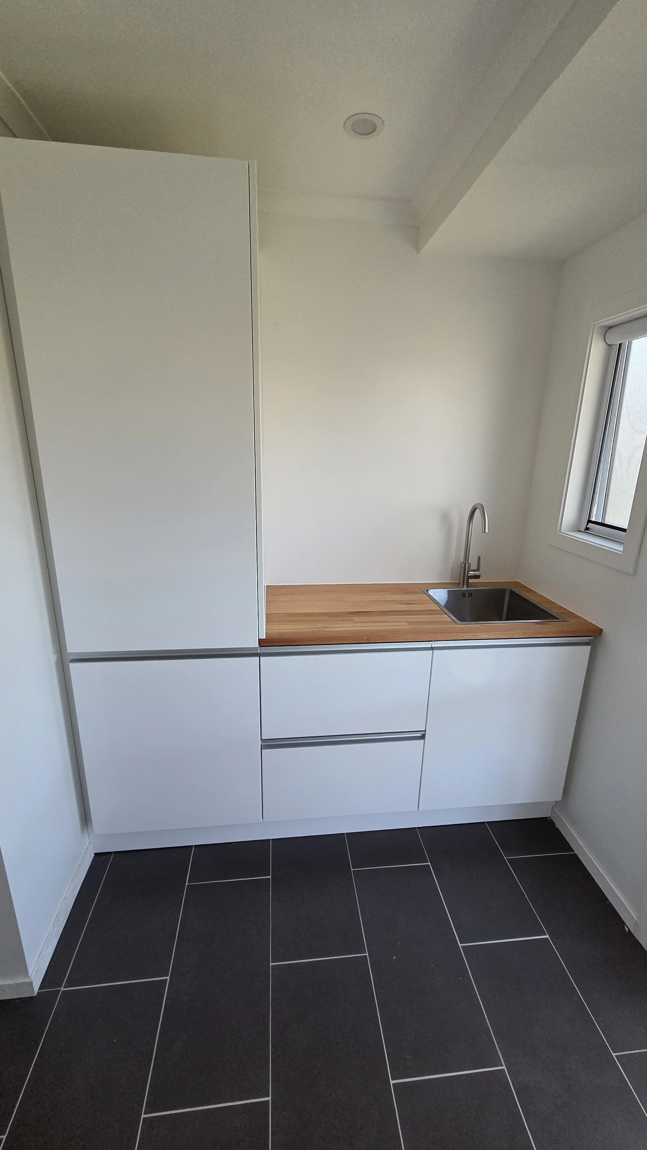 A small modern kitchen area with white cabinets, a wooden countertop, a stainless steel sink with a curved faucet, a window on the right, and black tiled flooring.