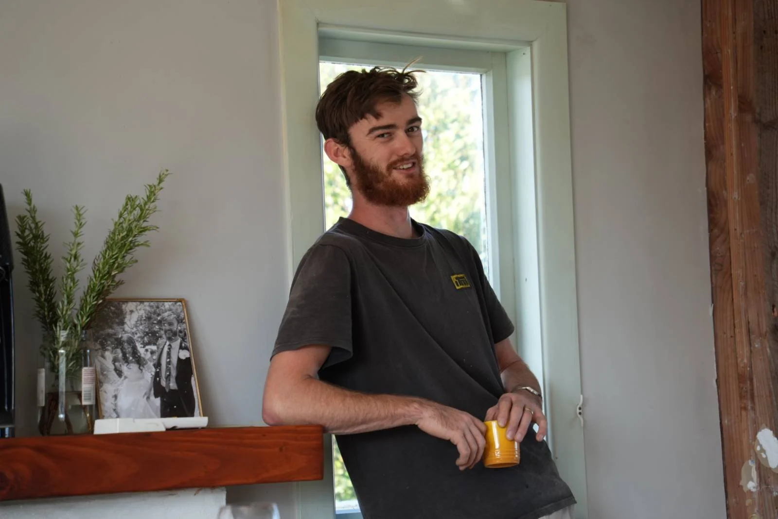 A young man with brown hair and a beard, wearing a black T-shirt, standing indoors near a window, holding a yellow mug, with a framed black-and-white photo and a small plant on a wooden surface behind him.