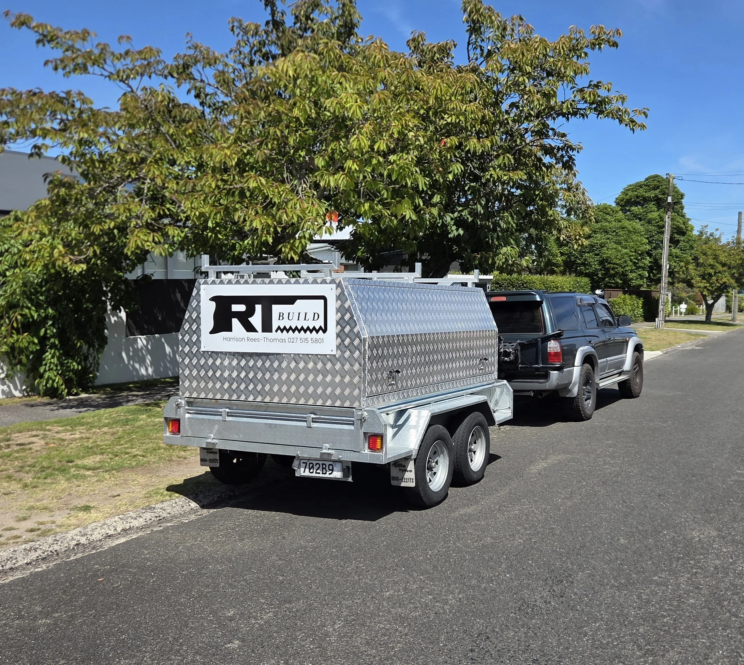 A utility trailer attached to a pickup truck parked in a residential neighborhood. The trailer has a sign that reads 'RBUILD' with contact details. In the background, there is a large tree with green leaves, a blue sky, and some houses and utility poles.