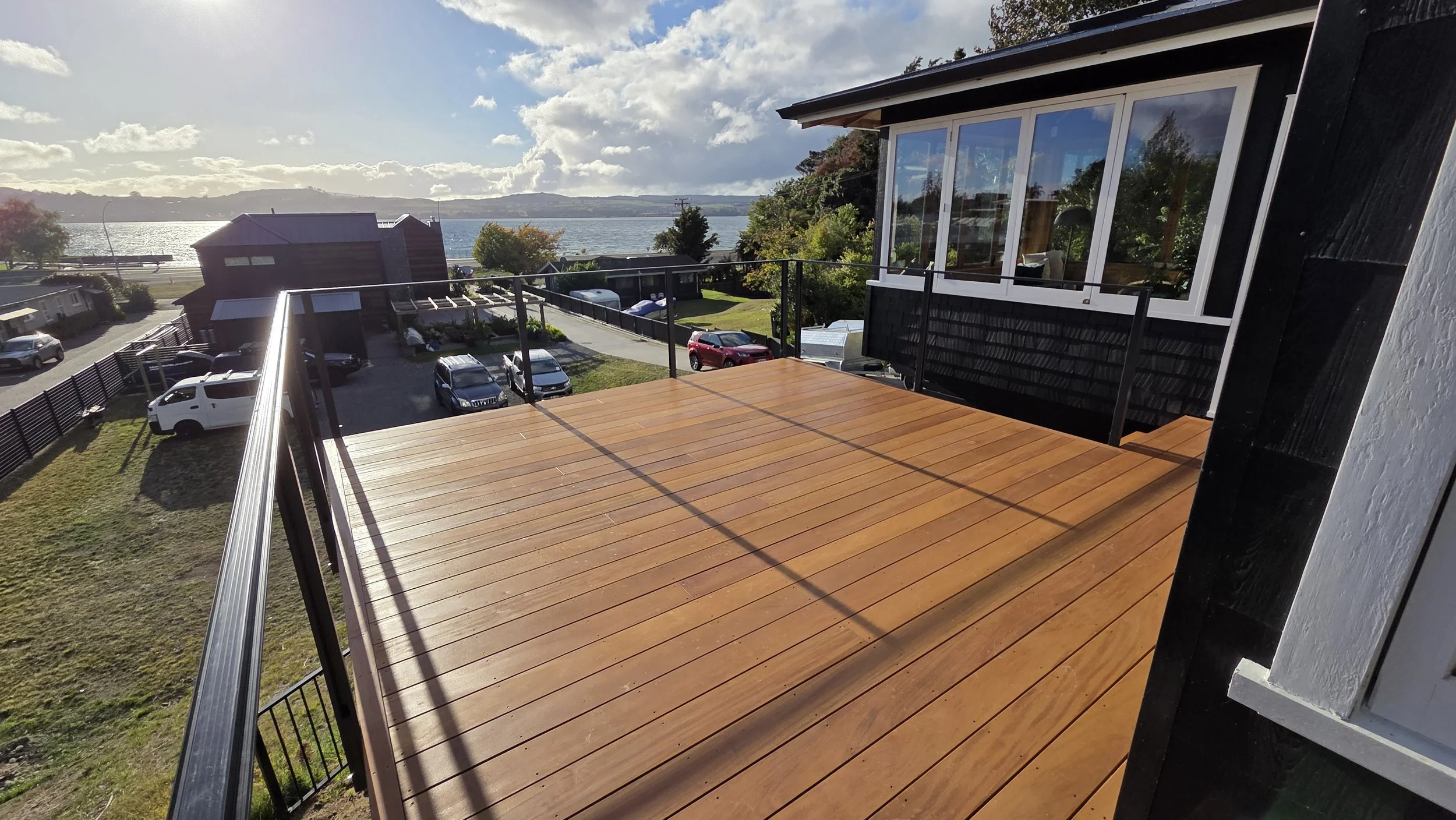 A wooden balcony overlooking a scenic view of a body of water, with cars parked below and a partly cloudy sky.