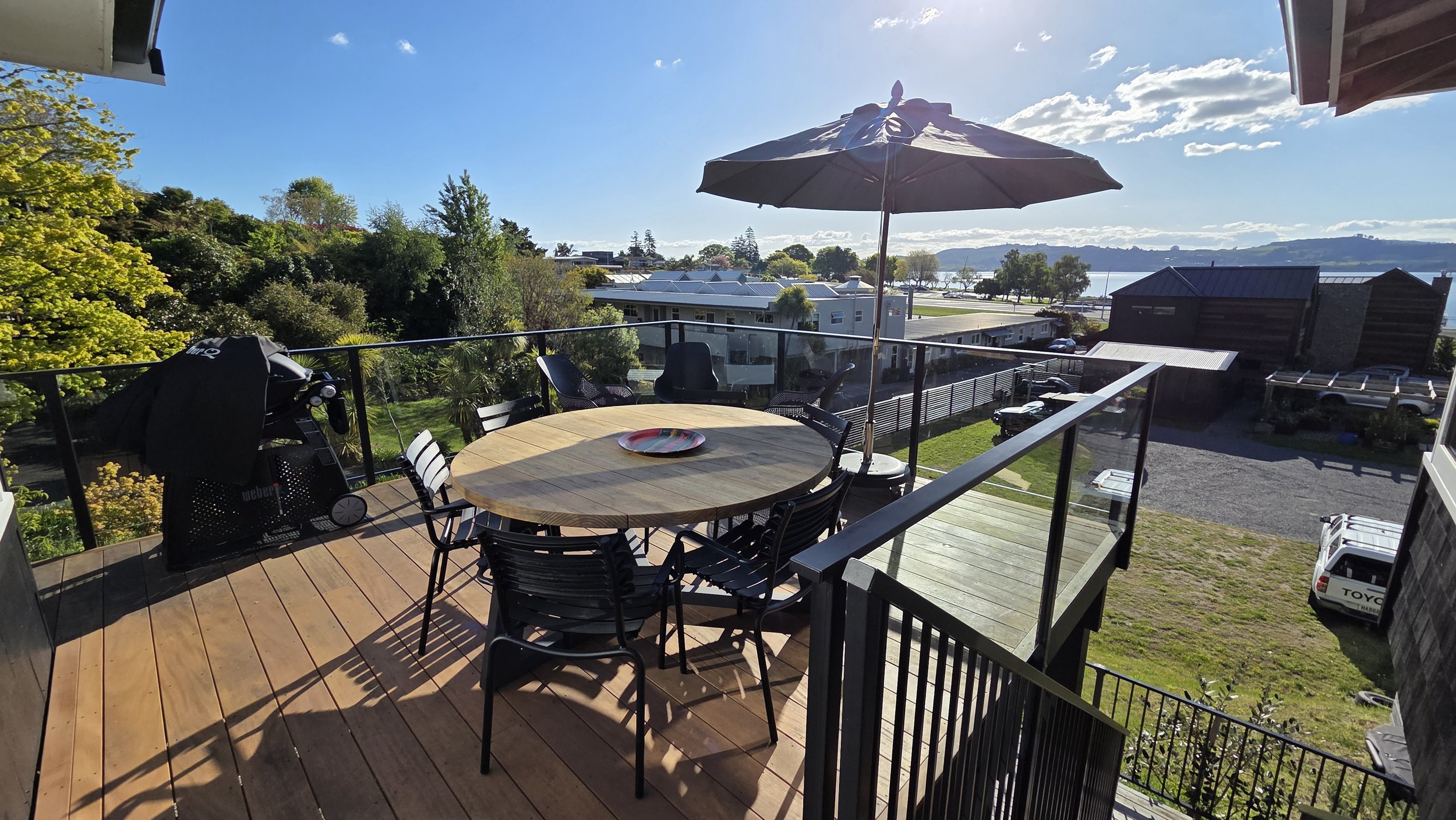 A balcony with a round wooden table, six black chairs, a large umbrella, a black barbecue grill, and a view of rooftops, trees, water, and a partly cloudy sky.