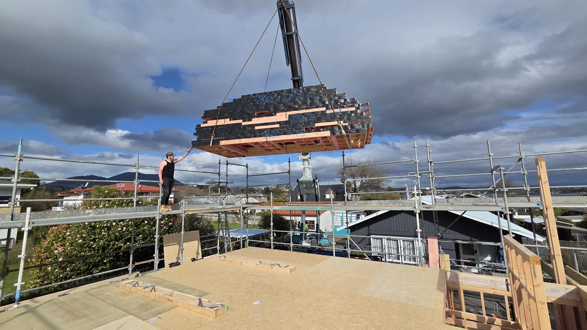 A construction worker stands on scaffolding near a house, guiding a large bundle of roofing materials being lifted by a crane against a cloudy sky.