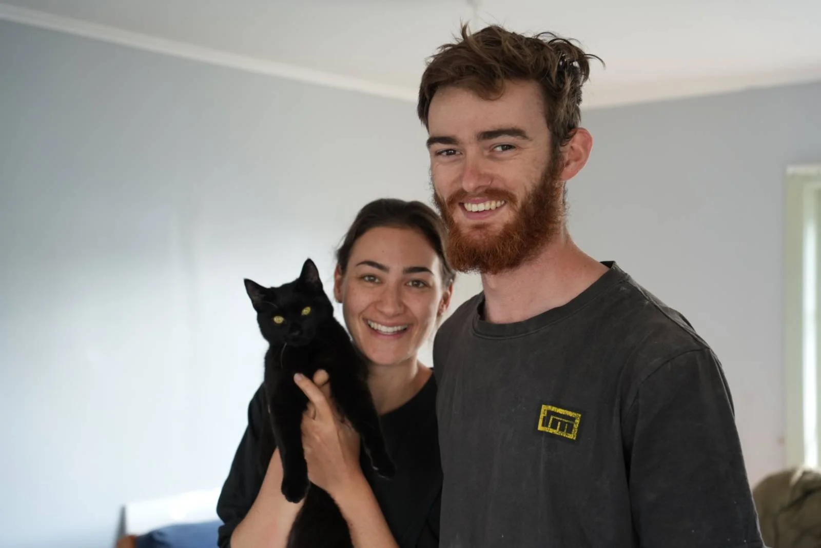 A smiling couple holding a black cat together, standing in a room with white walls and a window.