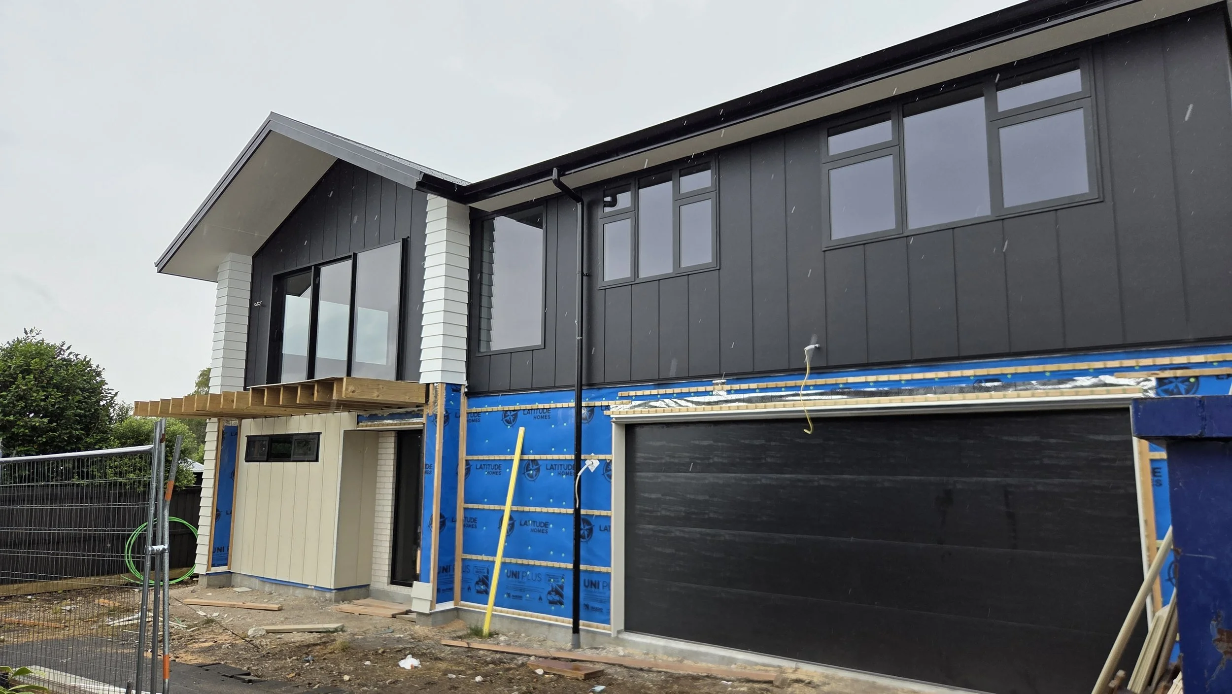 A two-story modern house under construction with black and white exterior walls. The upper floor has black siding with multiple windows, and the lower part has white siding. A wooden balcony or deck is being built on the front of the house. The construction site is fenced off with blue insulation on part of the ground floor, and tools and materials are visible around the area.