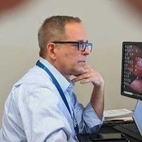 Man wearing glasses in a blue lanyard, sitting at a desk, looking at a computer monitor.