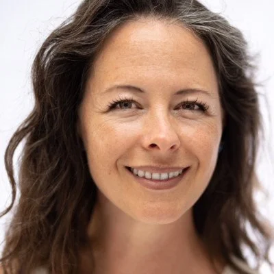 A woman with long, wavy brown hair smiling at the camera against a plain white background.
