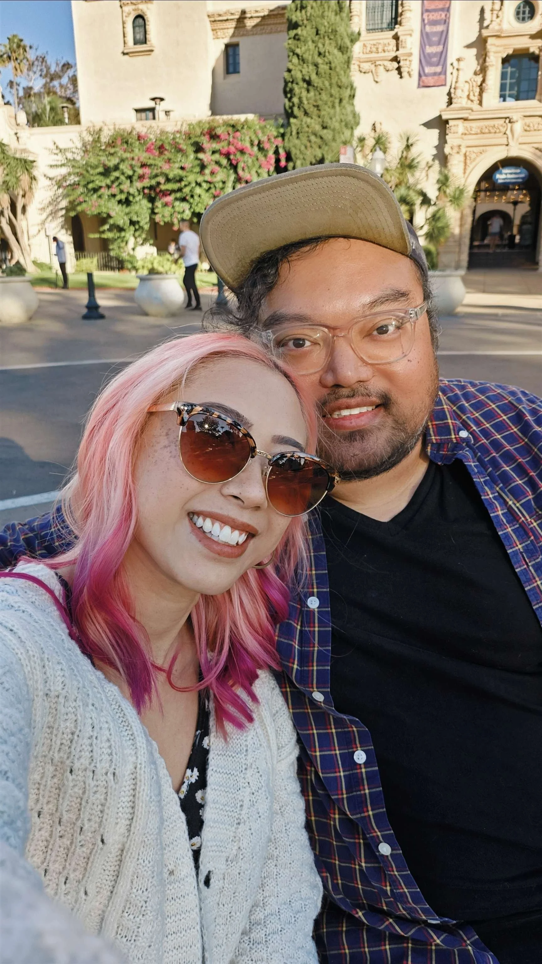 A smiling woman with pink hair and sunglasses taking a selfie with a smiling man wearing glasses and a baseball cap. They are outdoors with a historic building, trees, and flowering bushes in the background.