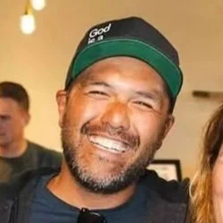 Man with a beard wearing a black cap that says 'God' smiling at a social gathering