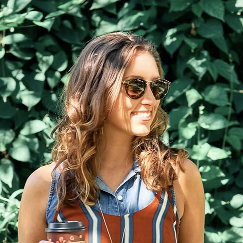 A woman with wavy brown hair wearing sunglasses and a sleeveless striped top, holding a coffee cup, standing in front of green leafy foliage.
