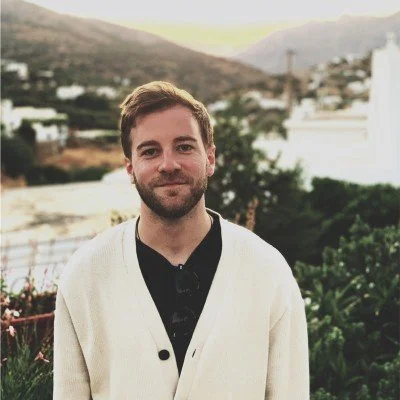 A young man with a beard and short brown hair standing outdoors in a hilly area with houses and trees in the background, wearing a light-colored cardigan over a dark shirt.