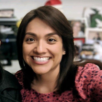 A woman with shoulder-length dark hair smiling in an indoor setting with blurred background and various objects.