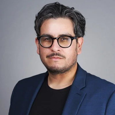 A man with dark hair, glasses, and a mustache, wearing a navy blazer and black shirt, posing against a gray background.