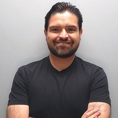 A man with dark hair and a beard, smiling, wearing a black t-shirt, standing against a plain gray wall.