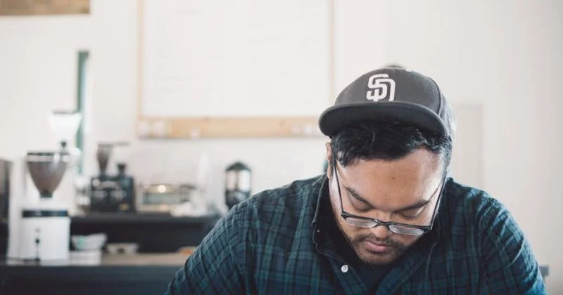 A man wearing a San Diego Padres baseball cap and glasses, looking down in a cafe or kitchen setting.