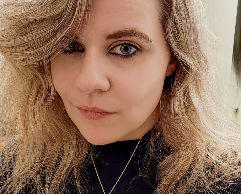 A woman with light blonde, wavy hair and blue eyes, wearing a black top and a gold necklace, looking at the camera.