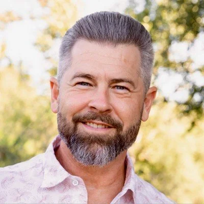 A smiling middle-aged man with short, dark hair and a beard, outdoors with trees in the background.