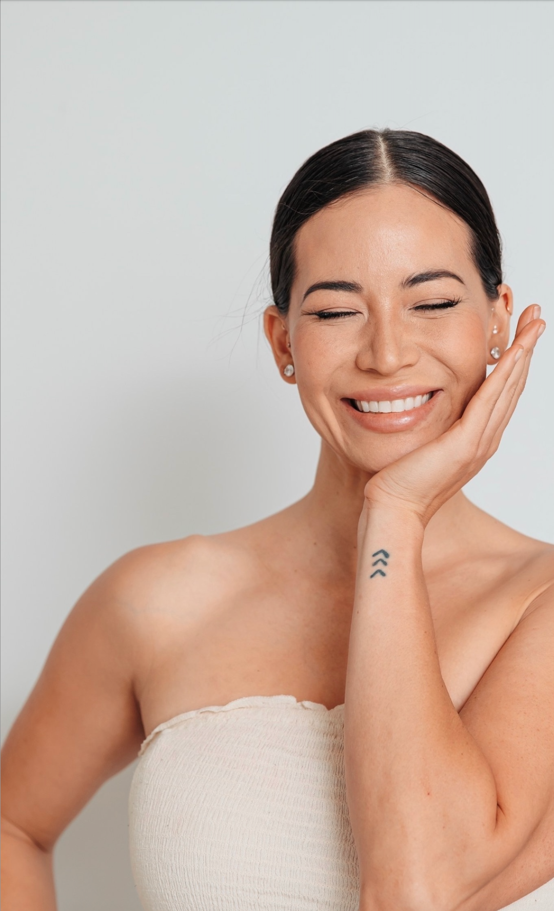 A smiling woman with dark hair pulled back, wearing pearl earrings and a strapless top, touching her face gently with her right hand which has a small tattoo of three upward arrows on her forearm.