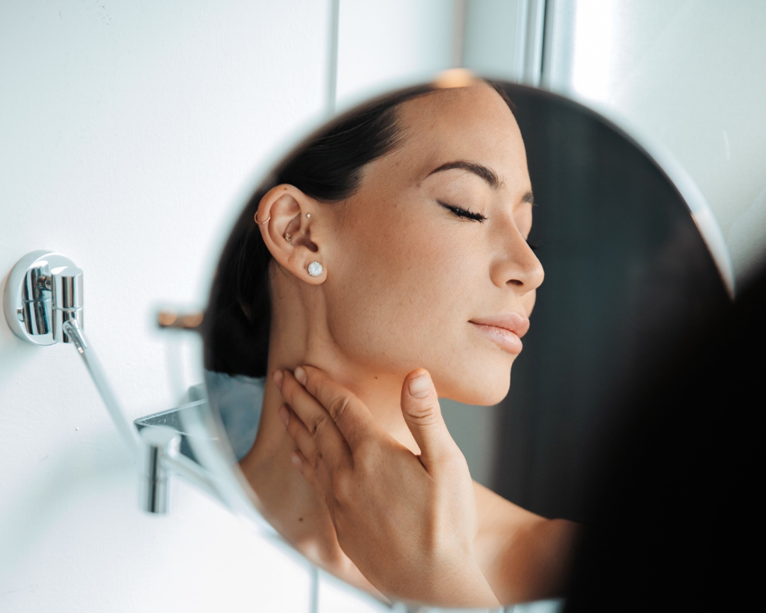 Close-up of a woman looking in a mirror, with her hand on her neck, wearing pearl earrings, with her eyes closed and a neutral expression.