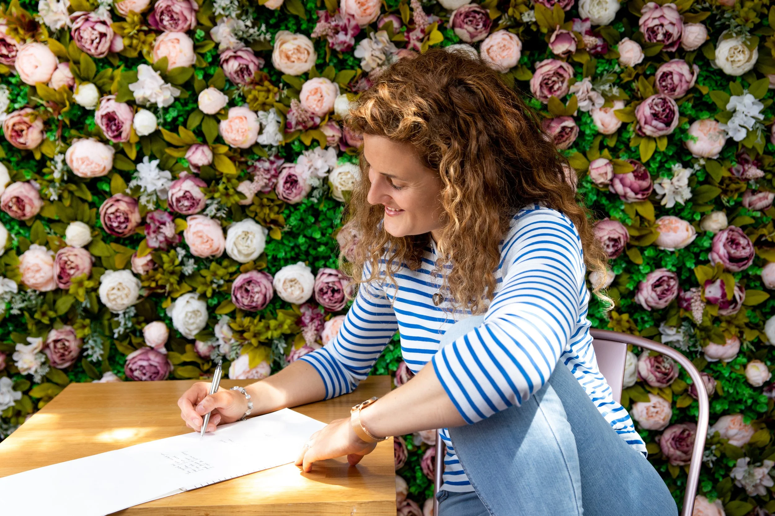 A woman with curly hair wearing a blue and white striped shirt, sitting at a wooden table, writing on a paper with a pen, in front of a colorful flower wall.