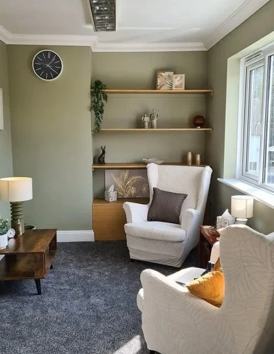 A cozy living room corner with beige armchairs, a wooden side table, and open shelves on a light green wall decorated with plants, books, and decorative objects. A window brings natural light into the room.