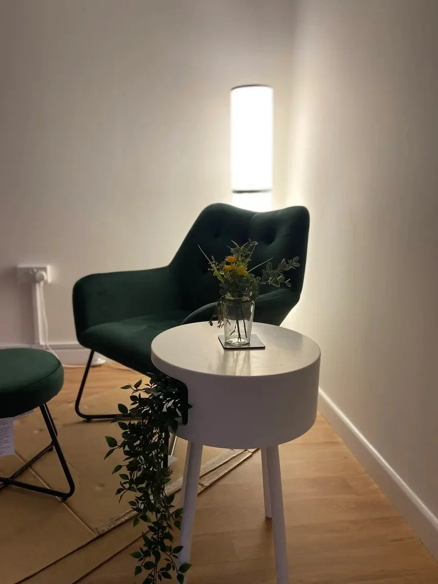 A cozy corner with a green armchair, a small white side table with a plant, and a tall floor lamp in a minimally decorated room.