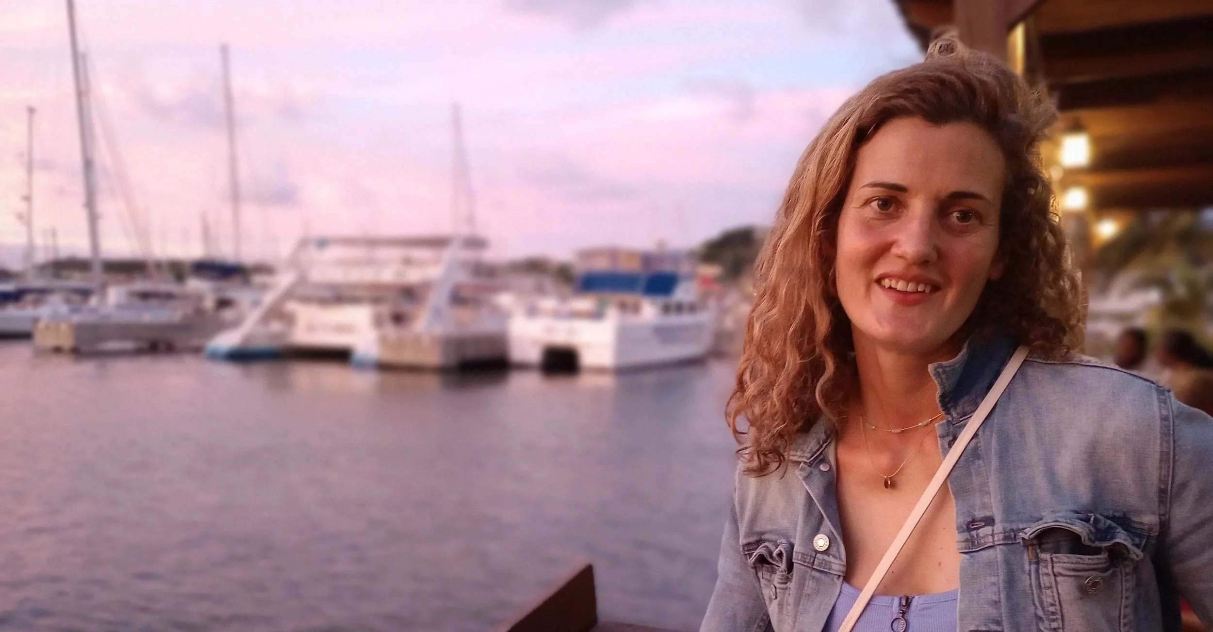 A woman with curly hair smiling at the camera on a dock with boats and a harbor in the background during sunset.