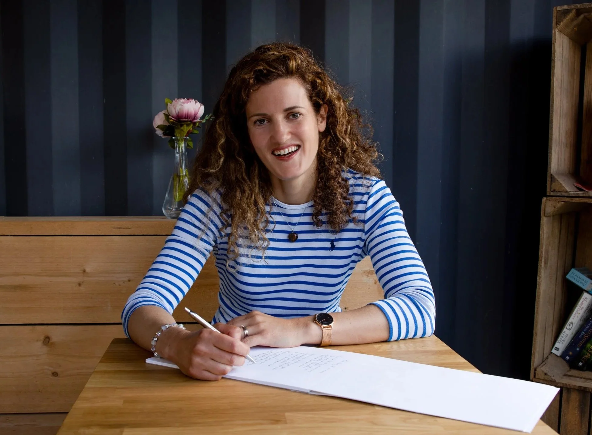 A young woman with curly brown hair, wearing a blue and white striped shirt, is smiling while writing on a piece of paper at a wooden table. There is a glass vase with pink flowers behind her on the table, and a dark wall with wooden shelving to her right.