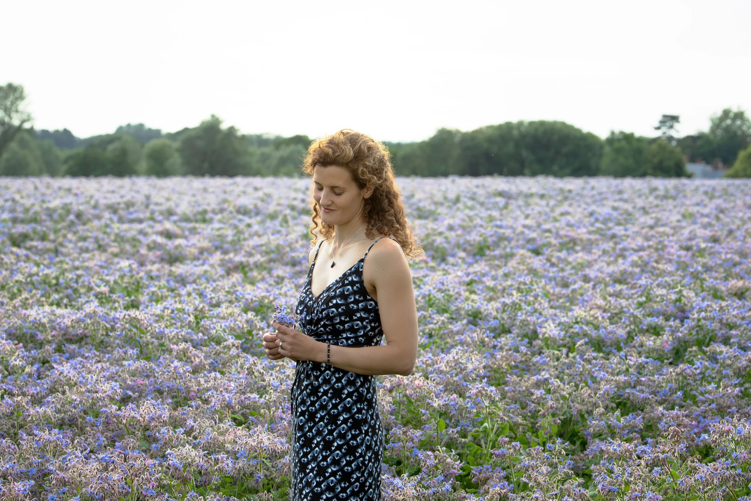 A woman with curly red hair in a black and white patterned dress standing in a field of purple and white flowers, holding some flowers in her hands, with trees and a partly cloudy sky in the background.