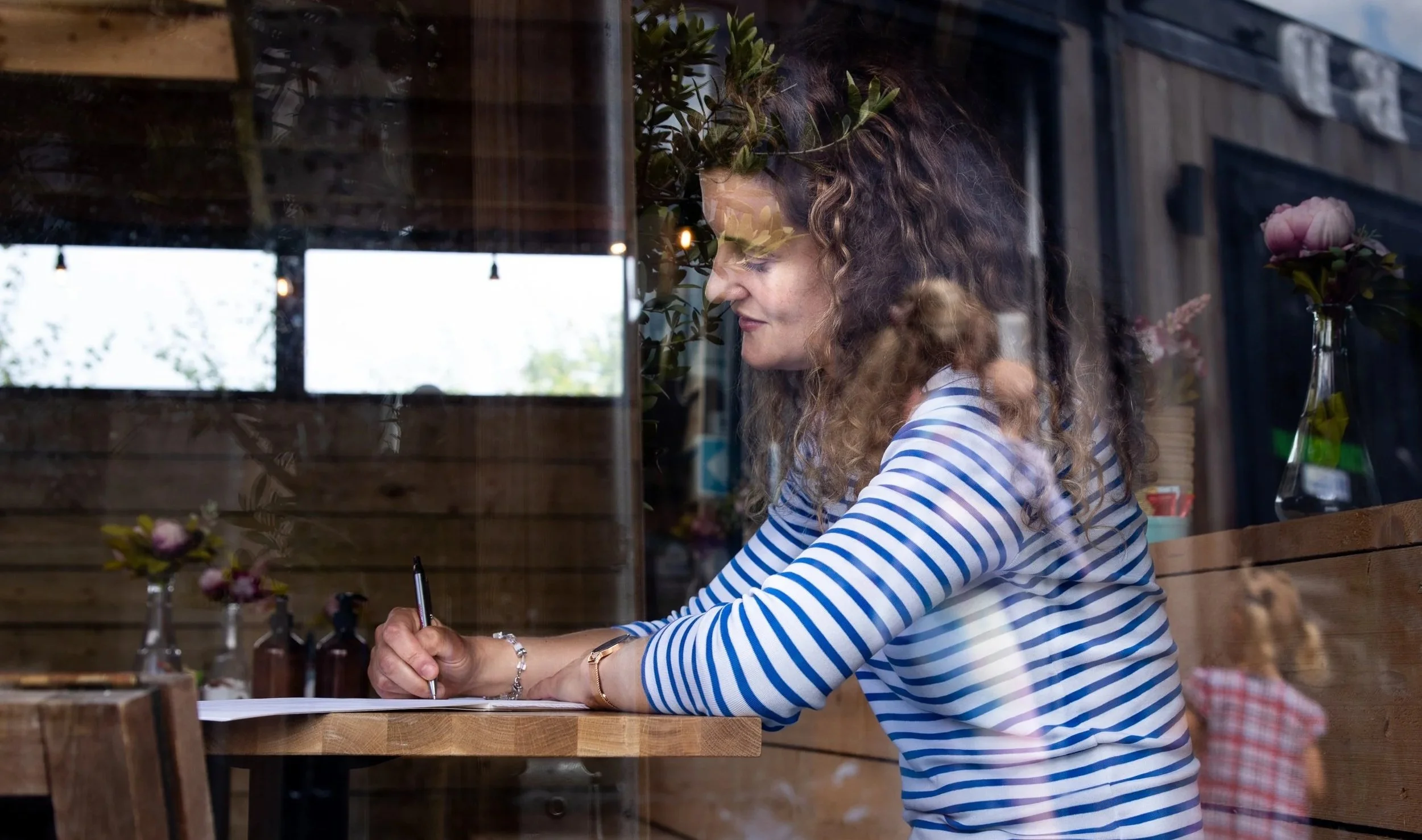 A woman with curly brown hair wearing a blue and white striped shirt, sitting at a wooden table inside a cafe, writing on paper with a pen, surrounded by flower vases and natural light coming through a nearby window.