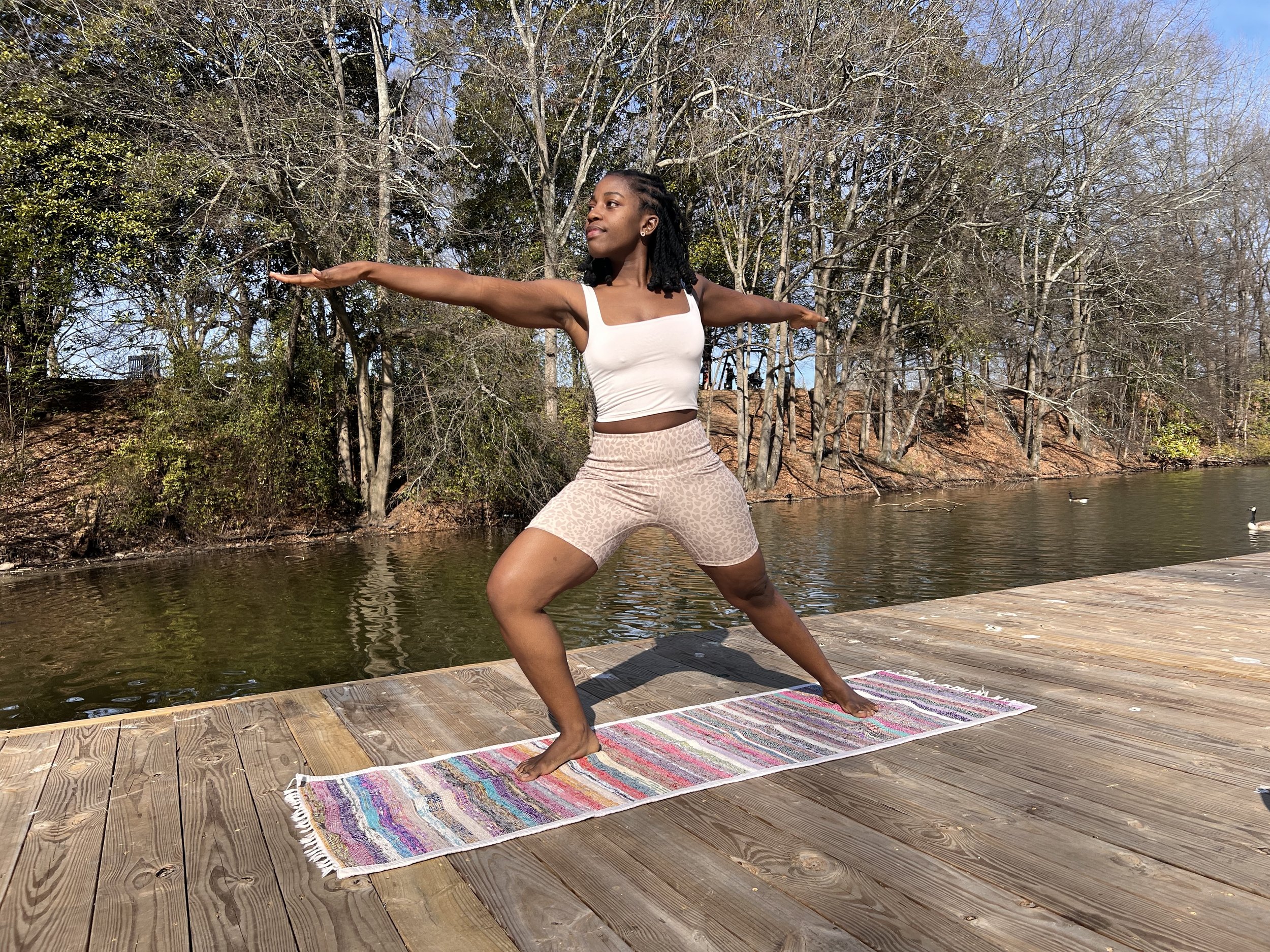 Young woman practicing yoga outside on a colorful mat near a lake, with trees and blue sky in the background.