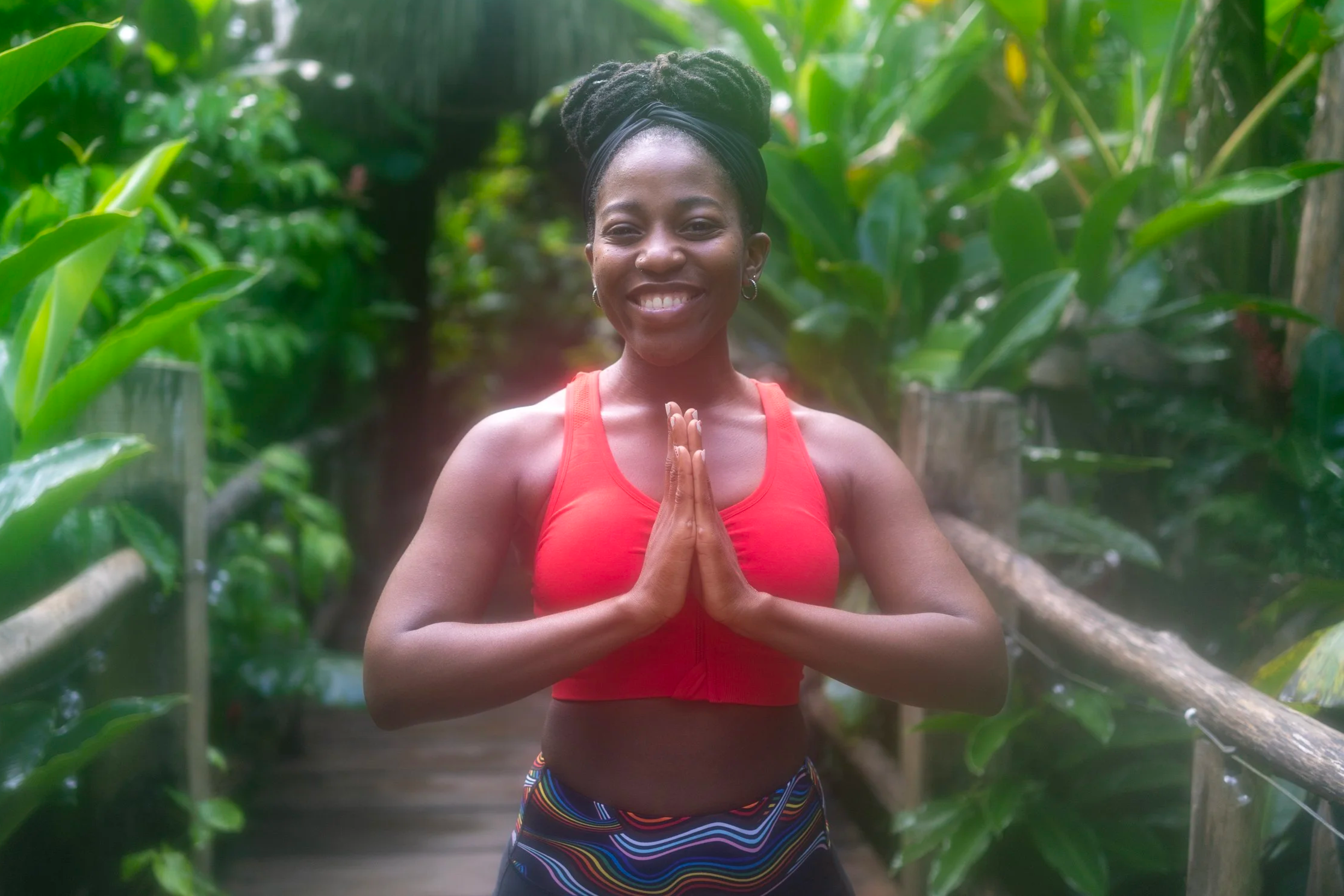 A woman practicing yoga outdoors among lush green plants, wearing a red tank top and colorful striped shorts, with hands pressed together in a prayer position.