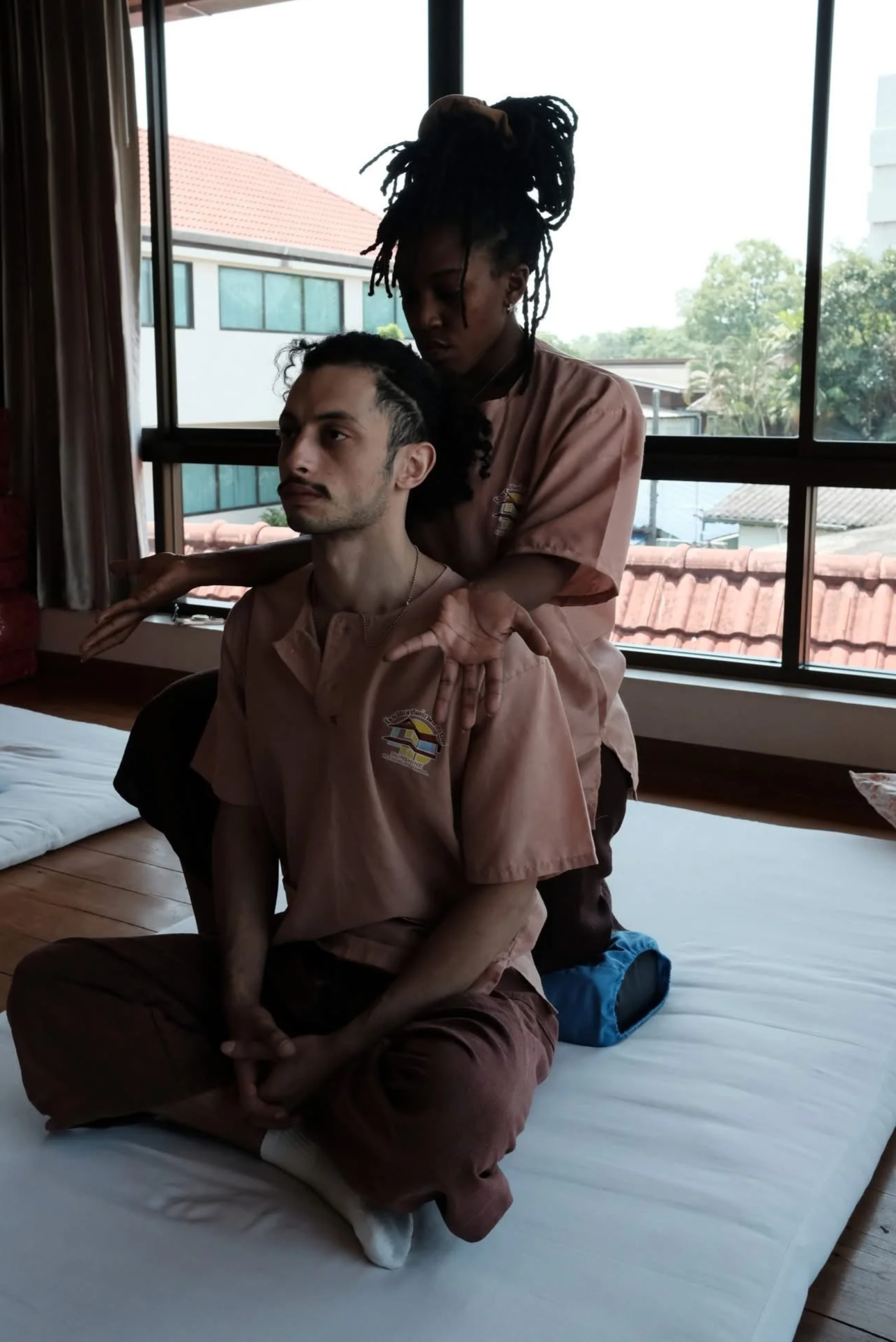 A person with dreadlocks giving a seated massage to another person with short straight hair in a room with large windows and a view of neighboring buildings and trees.