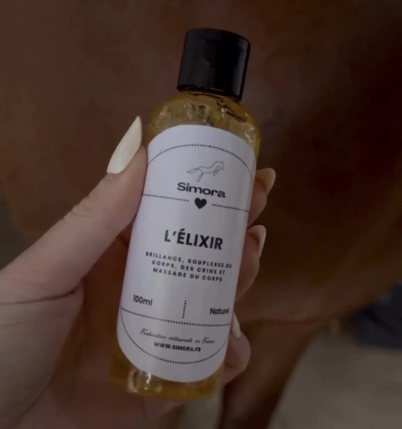 Une main tient une bouteille de 100 ml d'élixir nommé 'Simora'. Le flacon est transparent avec un liquide doré à l'intérieur. L'étiquette est blanche avec du texte en français, mentionnant des bienfaits tels que la brillance et la souplesse du corps,
