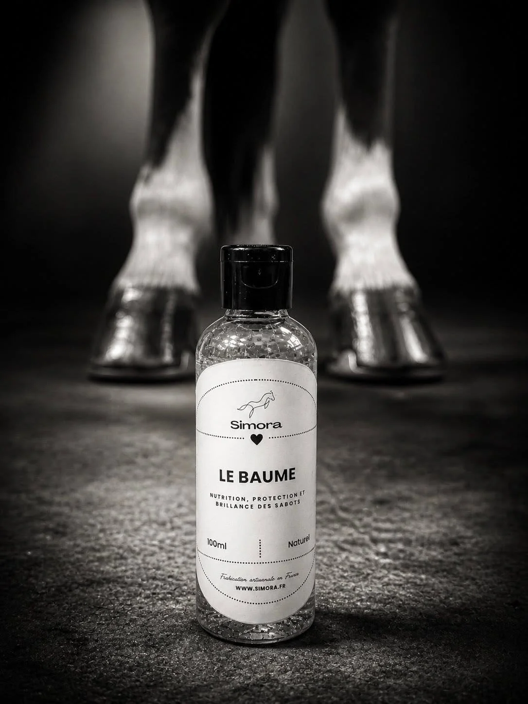 Une bouteille de lotion nommée 'Le Baume' de la marque Simora, avec un cheval figurant sur l'étiquette, posée sur un sol sombre, avec un cheval en silhouette en arrière-plan.