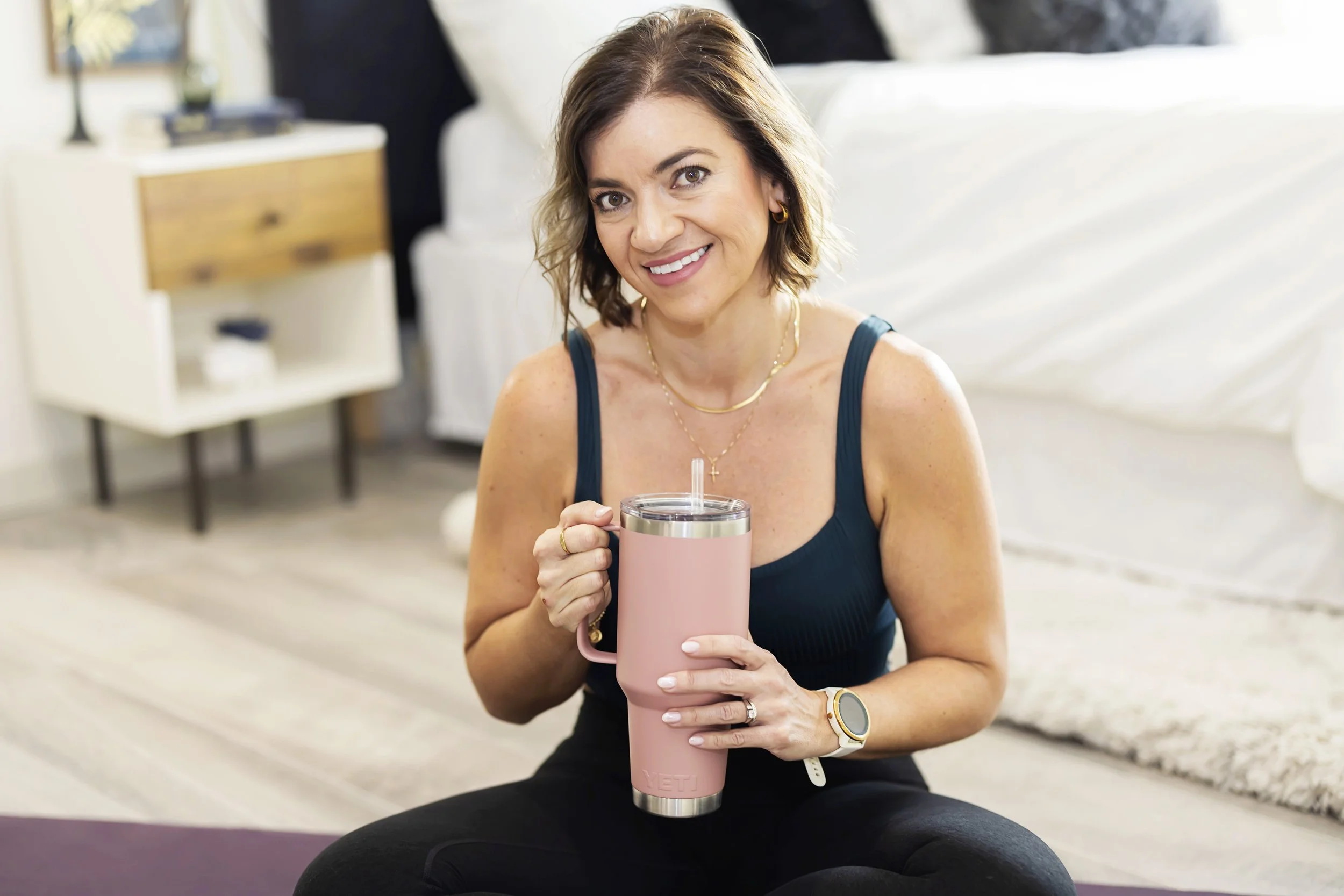 A woman sitting on the floor in a bright house, smiling and holding a pink YETI tumbler with a straw, wearing a navy blue tank top, gold jewelry, and a smartwatch.