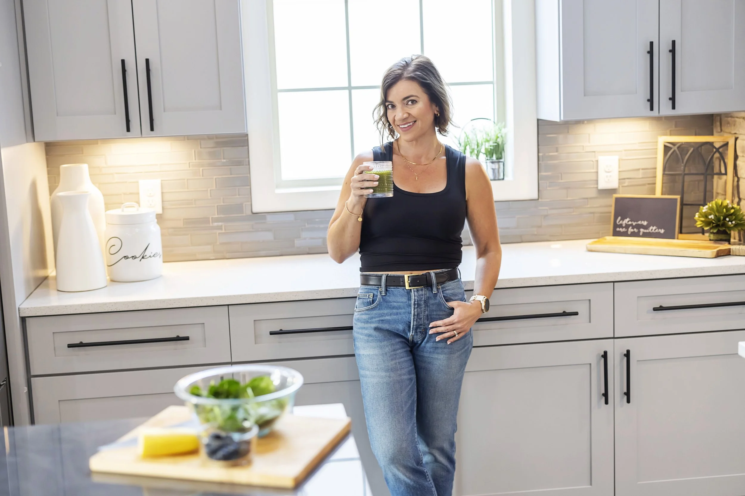 A woman with shoulder-length brown hair, wearing a black sleeveless top and blue jeans, is standing in a modern kitchen holding a glass of green smoothie, smiling at the camera.